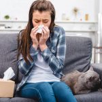 girl blowing nose in facial tissue while sitting with cat on couch